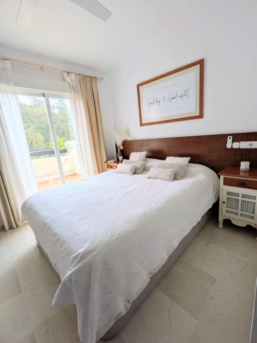 Luminoso dormitorio principal en BH Marbella con ropa de cama blanca, cabecero de madera y acceso a terraza con vistas al jardín