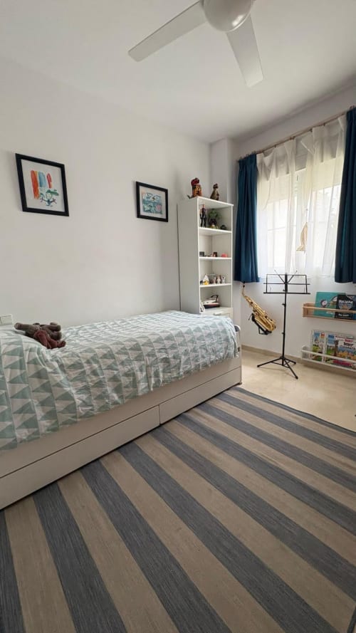 Habitación moderna y luminosa en BH Marbella con piso de rayas, cama blanca, estanterías y cortinas azules.