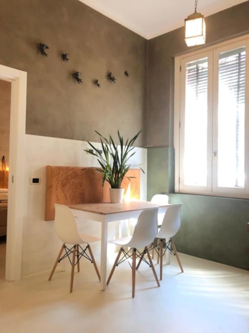 Zona pranzo moderna di Casa Città Brescia con sedie bianche, tavolo in legno, pianta in vaso e pareti beige con decorazione murale di uccelli.