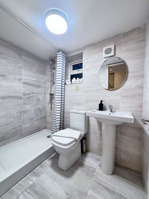 Modern bathroom with gray tile walls, white pedestal sink, toilet, round mirror, and shower in High Wycombe accommodation