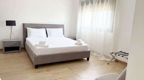 Dormitorio moderno en el hotel DakarByDays con cama tapizada gris, sábanas blancas, mesita de noche, lámpara y cortinas translúcidas en Dakar.