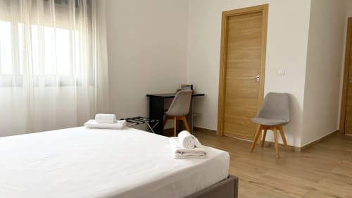 Modern bedroom in DakarByDays apartment with white bed, wooden desk, chair, and city view in Dakar, Scat Urbam