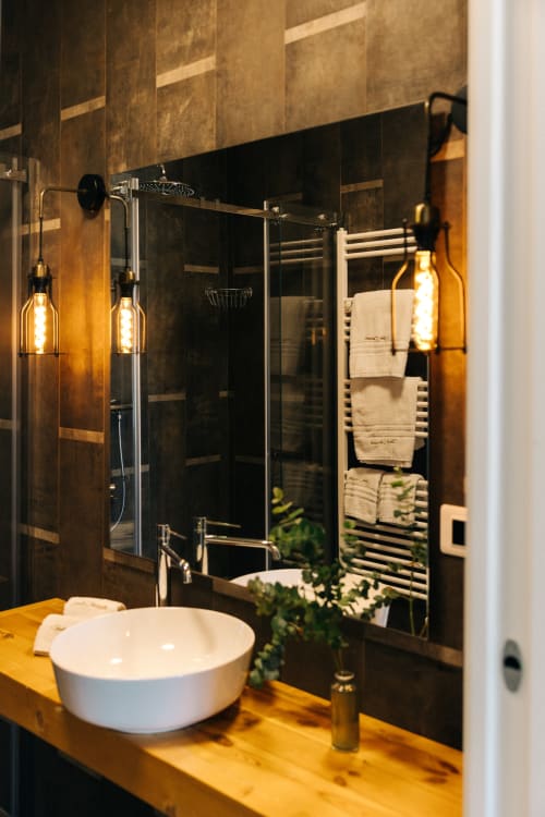 Moderno bagno presso Tenuta Fascianello con vanity in legno giallo, lavabo bianco, doccia con piastrelle scure e soffione pioggia, e