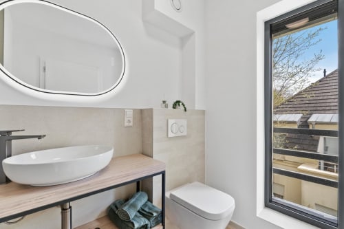 Salle de bain moderne à Mattbnb Metz avec miroir ovale, vasque blanche, toilettes et vue sur la ville par large fenêtre