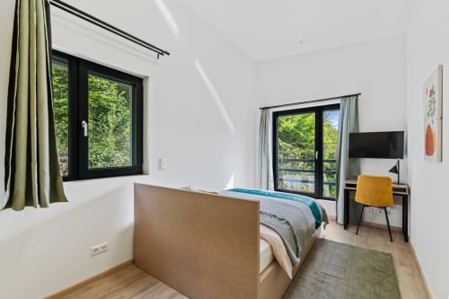 Chambre moderne à Mattbnb Metz avec lit, bureau, fauteuil jaune, TV écran plat et vue sur le jardin par fenêtres à cadre noir