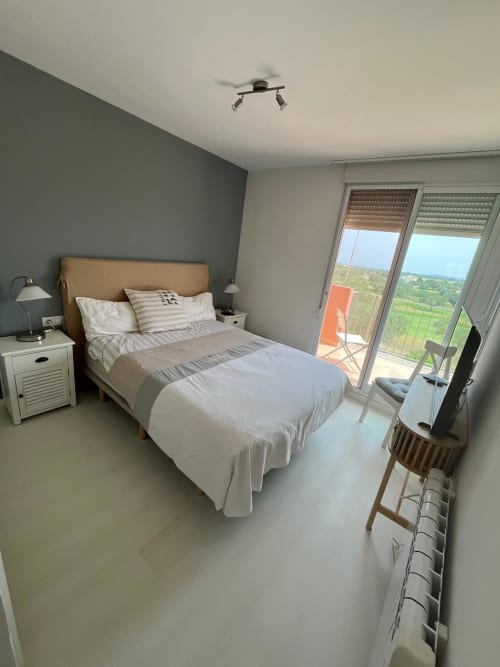 Dormitorio moderno en apartamento Sant Jordi con vistas al golf, cabecero en tonos beige, pared gris y balcón con vista al paisaje verde