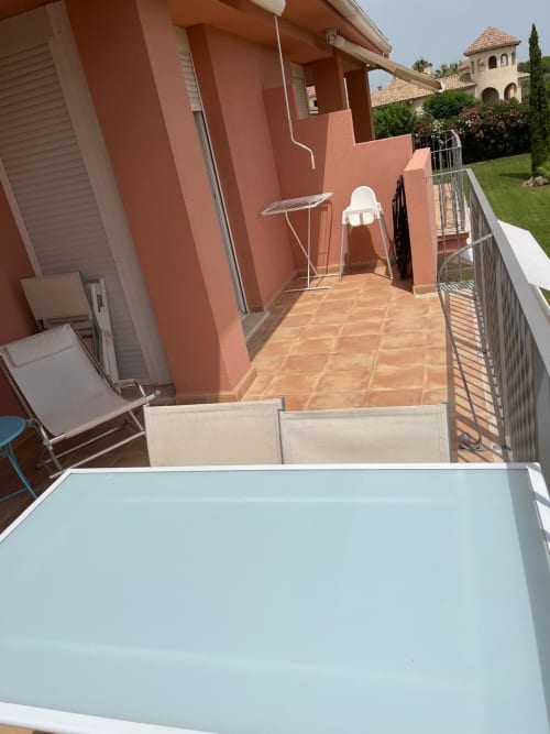 Terraza soleada del apartamento Sant Jordi con mesa, tumbonas y edificio de muros rojos con vistas a los jardines del campo de golf