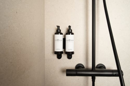 Amenities de baño en Hotel Bruna: dos botellas dispensadoras y accesorios de ducha negros sobre pared beige