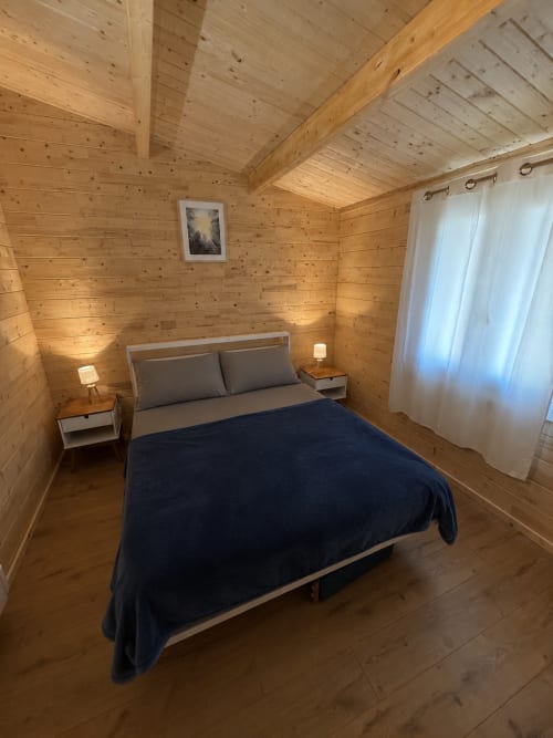 Chambre douillette de lodge en bois avec lit queen-size, oreillers gris, couverture bleu foncé, tables de nuit en bois avec lampes, rideaux blancs et