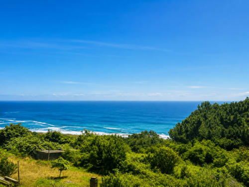 Vista costera del hotel Holibai en Baiona, España con océano azul, playa y exuberante vegetación verde