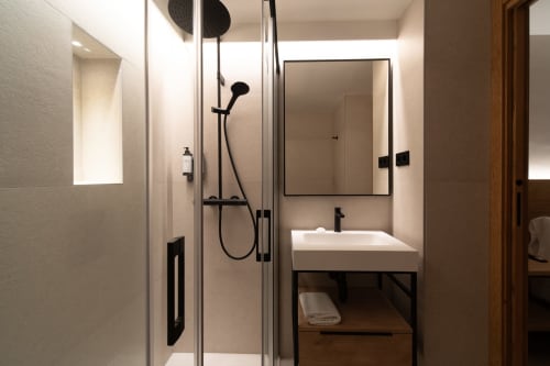 Moderno baño del Hotel Bruna con ducha a ras de suelo, grifería negra, lavamanos blanco y espejo iluminado en Esterri D'Àneu