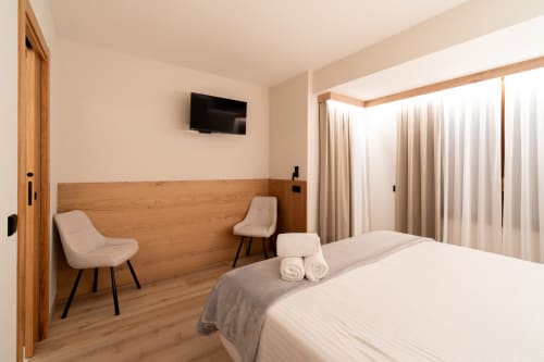 Dormitorio doble del Hotel Bruna en Esterri D'Àneu con cabecero de madera, sillas modernas, TV y grandes ventanas con cortinas