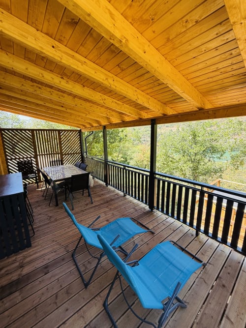 Terrasse en bois aux Cabanes du Belvédère avec chaises longues bleues, espace repas, vue sur lac et plafond à poutres apparentes
