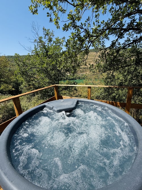 Bain à remous fumant sur terrasse en bois dominant la rivière Ain et les collines boisées du gîte Cabanes du Belvédère