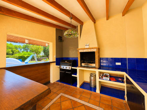 Casa Emilia villa kitchen with pool view, yellow walls, blue tile accents, fireplace, and outdoor terrace in Moraira