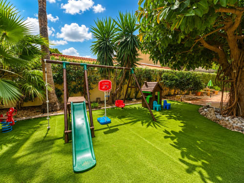 Sunny children's play area at Casa Emilia villa in Moraira with slide, swing, basketball hoop, and tropical garden