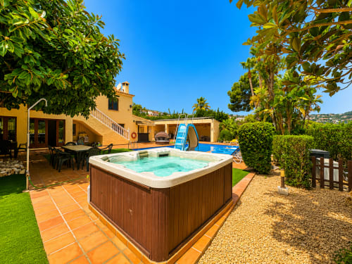 Moraira villa Casa Emilia backyard with hot tub, swimming pool, yellow house and lush greenery under blue sky