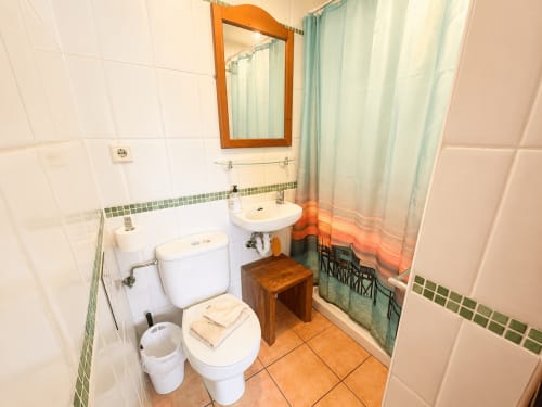 Clean bathroom with white tiles, wooden mirror, toilet, sink, and shower with green curtain at Casa Emilia villa in Moraira