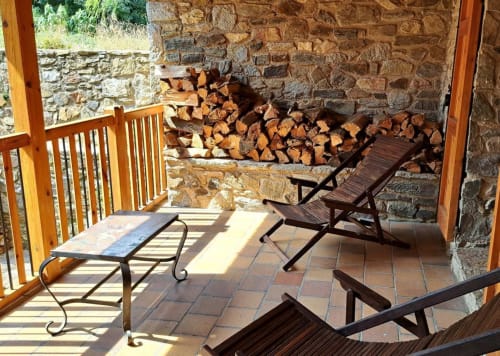 Patio cubierto en L'Orri Rectoria Nova de Campelles con muros de piedra, barandillas de madera, tumbonas y leña apilada.
