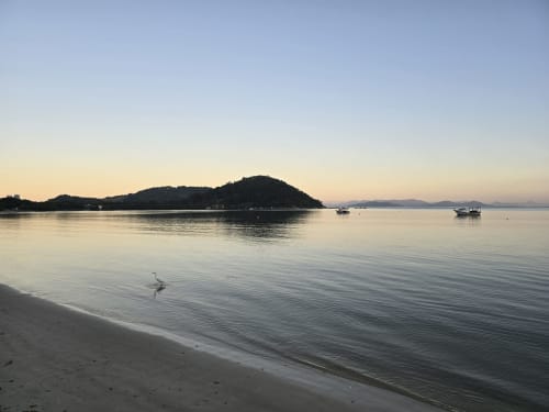 Vista serena da praia do hotel Philia em Florianópolis com águas calmas, barcos ancorados e ilha florestada ao amanhecer