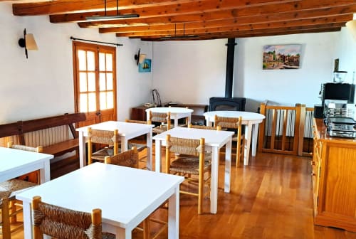 Comedor rústico en Campelles con vigas de madera, mesas blancas, sillas de paja y vistas a la montaña en L'Orri Rectoria