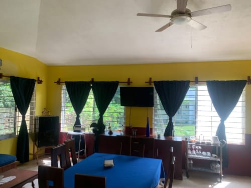 Villa de tonos amarillo brillante con comedor, mantel azul, muebles de madera oscura, cortinas negras, ventilador de techo y vistas al jardín
