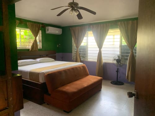 Dormitorio de la villa Casa Leticia con paredes verdes, cama queen, asientos naranjas, ventilador de techo y vistas al jardín a través de puertas con cortinas