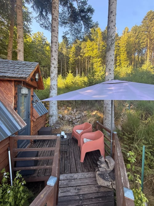 Terrasse de la Cabane L'Écureuil avec parasol bleu, chaises corail et vue sur la forêt aux Cabanes des Marchous, Granges-Aumontzey