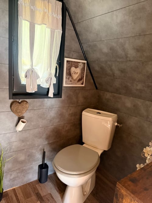 Salle de bains rustique du gîte Cabane L'Écureuil avec toilettes, décor bois, ornements cœur, fenêtre rideaux et montagne