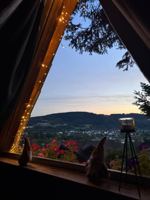 Vue sur la vallée au coucher de soleil depuis la fenêtre du gîte avec fleurs en pots, guirlandes lumineuses et paysage montagneux aux Cabanes des