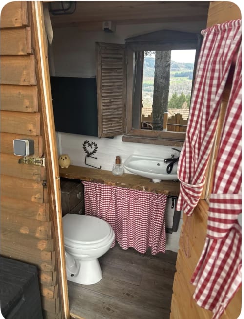 Salle de bain compacte de la Cabane Chouette aux Cabanes des Marchous avec toilettes, lavabo, rideaux à carreaux rouges et