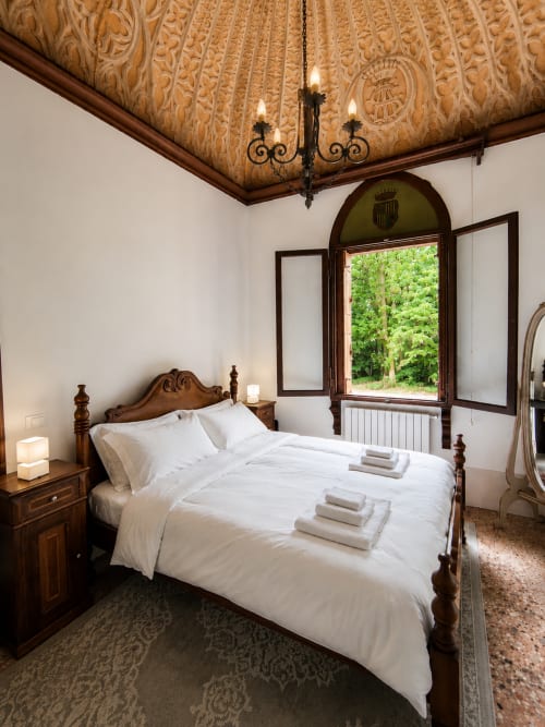 Elegant bedroom at Veneto Villa with carved wooden ceiling, four-poster bed, and arched window overlooking lake in