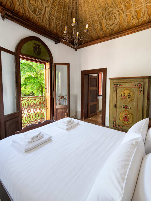 Elegant bedroom at Veneto Villa with white bed, ornate wooden ceiling, and balcony overlooking lush lake views in