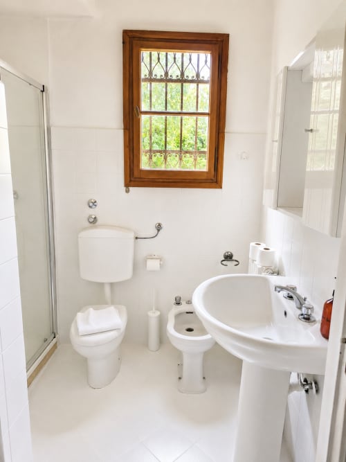 Clean white bathroom with toilet, bidet, pedestal sink, shower and wooden-framed window at Veneto Villa, Sant'Agostino