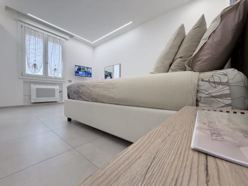 Camera da letto deluxe moderna Dream Suite Bologna con letto color crema, cuscini, comodino in legno, TV, finestra e