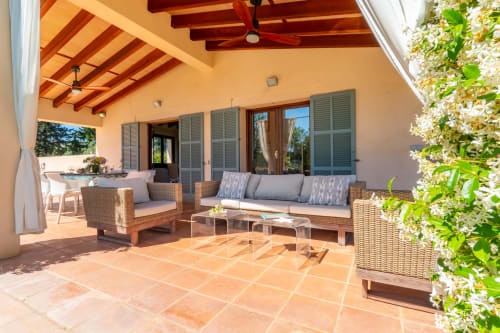 Terraza cubierta de la villa Can Mantellé en Artà con asientos de mimbre, vigas de madera y plantas en flor