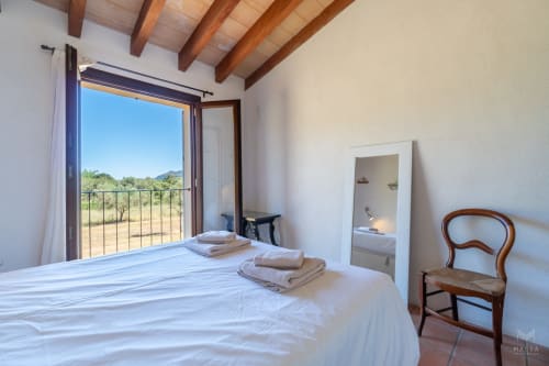 Dormitorio luminoso en Massa Properties de Artà con vigas de terracota, vista al balcón, espejo y silla de madera