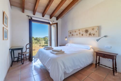 Dormitorio luminoso con cama blanca, vigas de madera y balcón con vistas al paisaje de Artà en Massa Properties