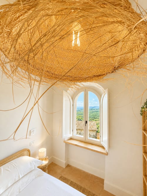Tuscan bedroom with arched window overlooking countryside, woven ceiling pendant, and white bedding at Montemassi guesthouse