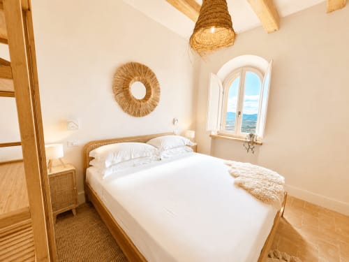 Bright Tuscan bedroom with wooden bed, white linens, woven accents, and panoramic countryside window at Montemassi retreat