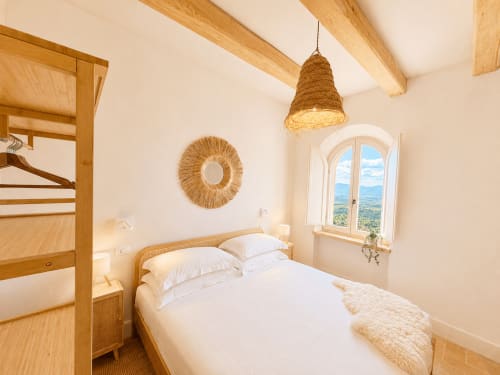 Bright Tuscan bedroom with wooden beams, arched window overlooking countryside, white linens, woven pendant light, and rattan