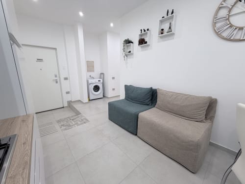 Monolocale moderno a Dream Suite Bologna con divano letto, lavatrice e pavimenti in piastrelle bianche in layout open-plan.