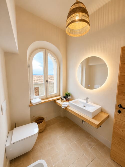 Modern bathroom in Tuscan Townhouse Bellavista Suite, Montemassi, featuring arched window, circular mirror, wooden vanity and