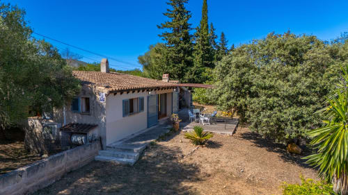 Encantadora casa de pueblo encalada en Artà con tejado de barro, rodeada de vegetación y árboles mediterráneos bajo cielo azul