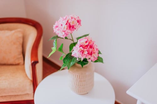Flores rosa de hortênsia em vaso creme sobre mesa branca ao lado de cadeira vermelha no quarto Villa Amore