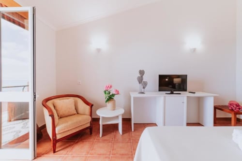 Quarto duplo com vista para o mar na Villa Amore, com piso em azulejo terracota, poltrona pêssego, escrivaninha branca e TV de tela plana