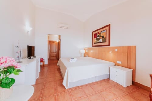 Espaçoso quarto de casal na Villa Amore com ladrilhos em terracota, cabeceira de madeira, roupa de cama branca, TV e flores rosa na escrivaninha