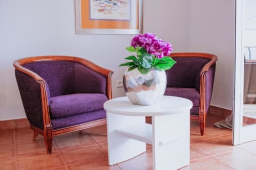 Poltronas estofadas em roxo com estrutura de madeira ao lado de uma mesa redonda branca com flores rosas na área de estar da Villa Amore