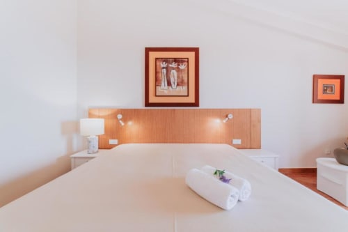 Quarto de casal moderno na Villa Amore com vista para o mar, cama branca, toalhas enroladas, cabeceira de madeira com iluminação e quadro