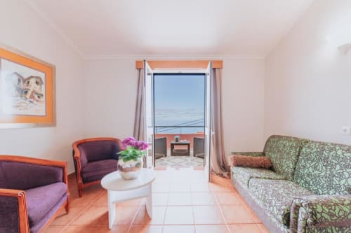 Sala de estar luminosa com vista para o oceano, poltronas roxas, sofá verde, mesa branca com flores e acesso à varanda da Villa